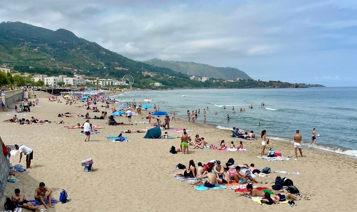 People relax and swim at a sandy beach with green hills along the coastline.