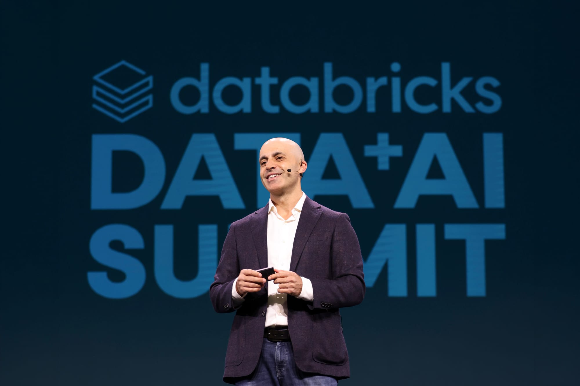 Databricks CEO Ali Ghodsi standing in front of Databricks Data and AI Summit logo.
