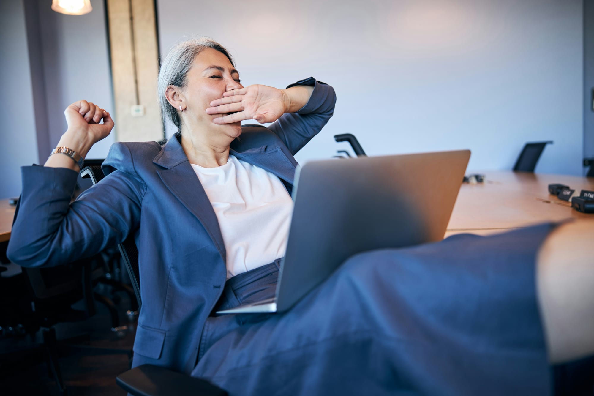 Woman in blue business suit with laptop on her lap, feet up, yawning.