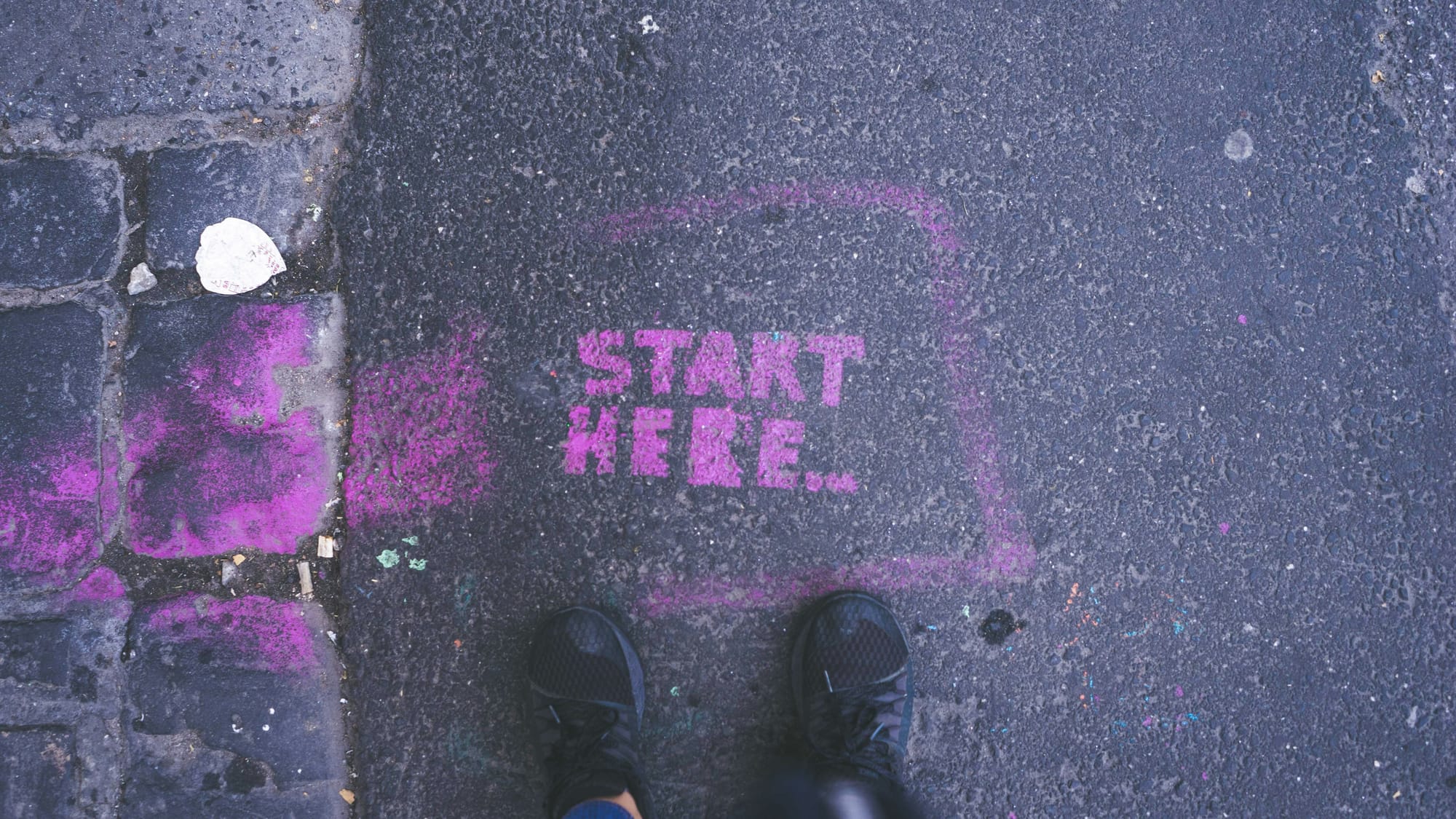 Pink "Start here" stenciled on sidewalk with a person with black shoes standing near it.
