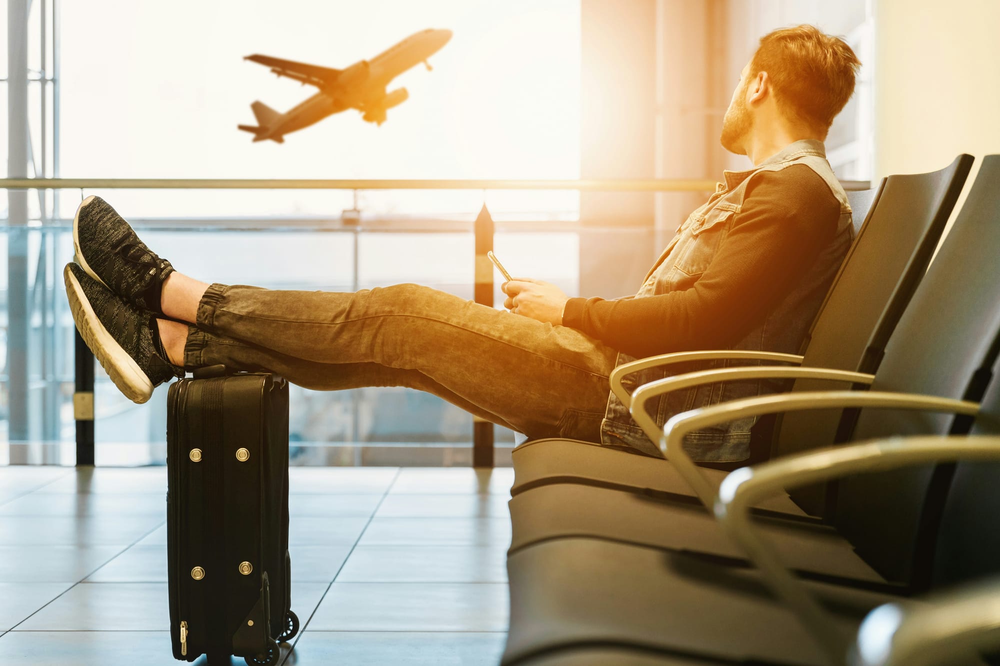 Man with feet up on suitcase at airport with plane taking off in background.