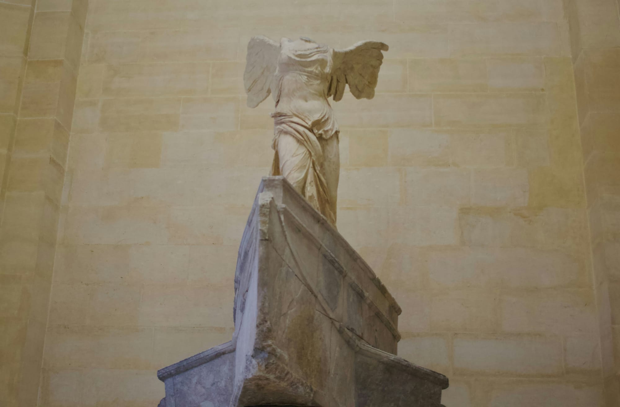Headless winged marble statue standing atop a large stone pedestal.