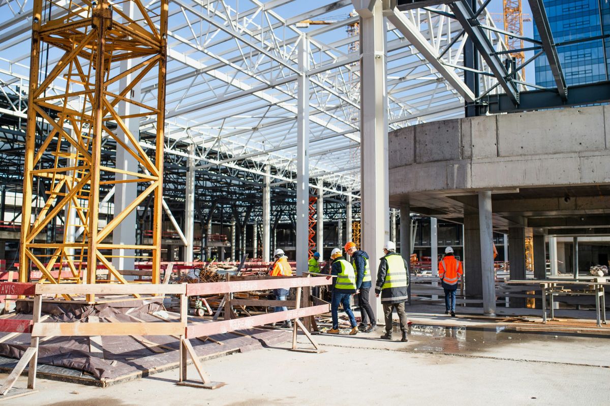 Construction workers wearing safety vests and helmets walk through a large steel-framed building under construction, symboliz