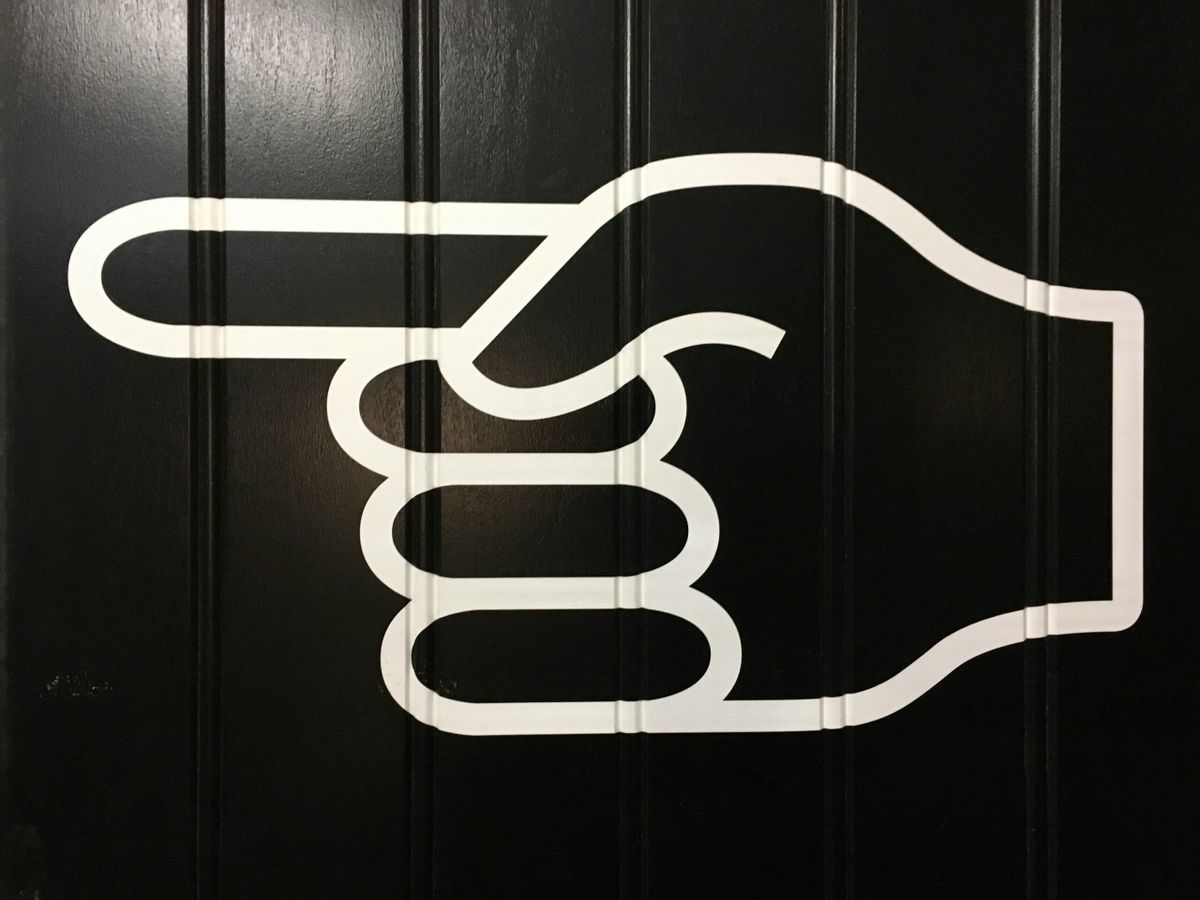 A large white graphic of a hand with the index finger pointing left, displayed on a black paneled wall.