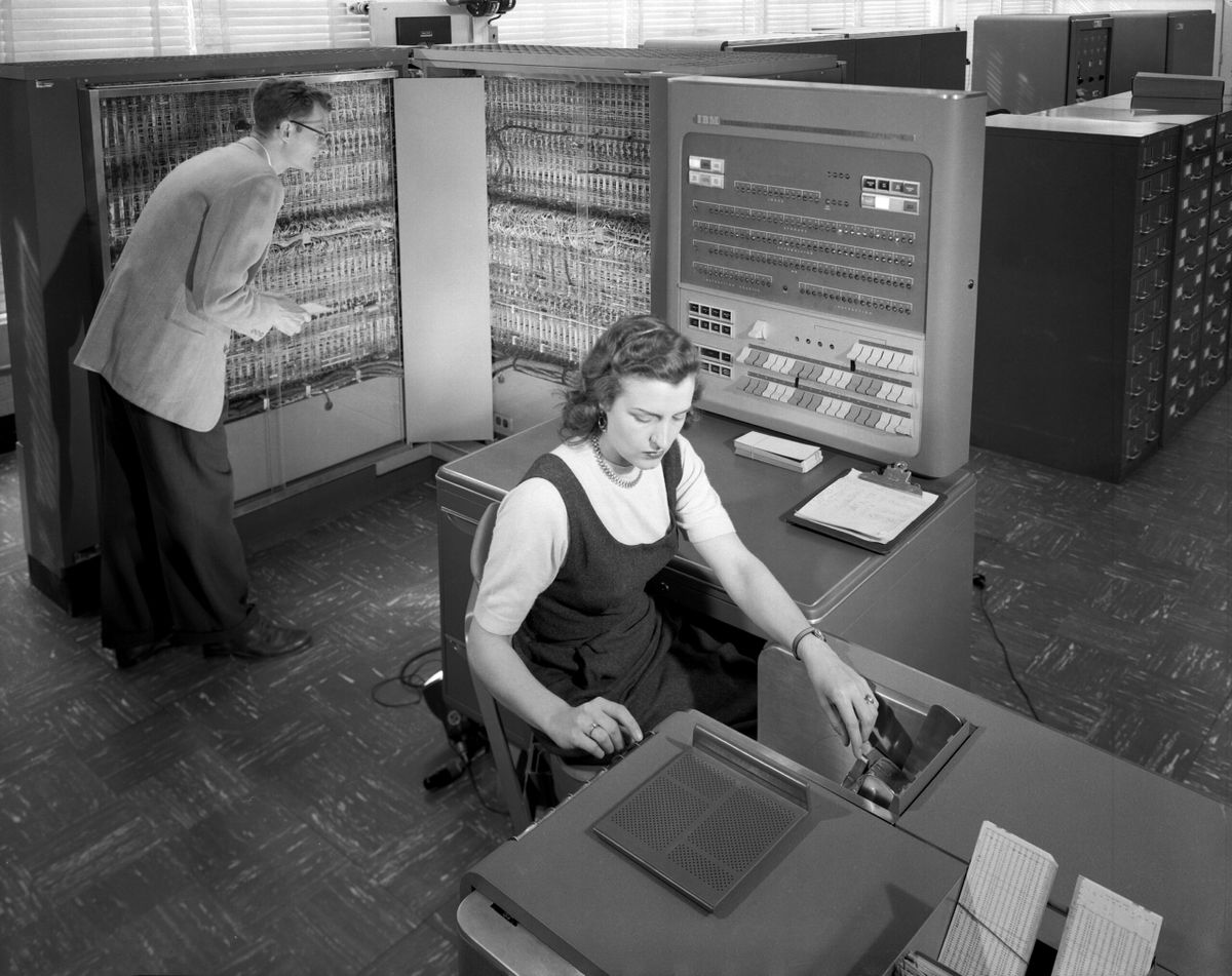 IBM mainframe from the 1950s or 60s. A woman is sitting in front of a huge keyboard for data entry. A man is looking inside o