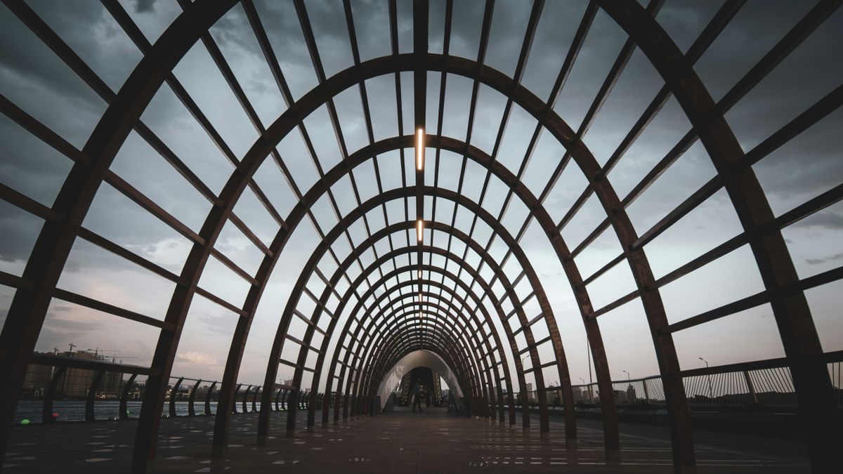 A wide-angle view of a modern arched walkway made of evenly spaced metal beams forming a tunnel-like structure, with a cloudy