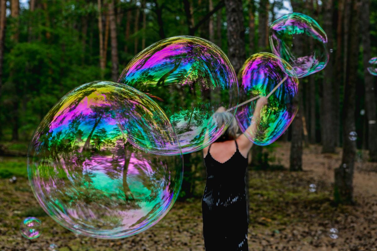 A person in a black dress creating giant iridescent soap bubbles in a forest clearing, with trees reflecting in the colorful