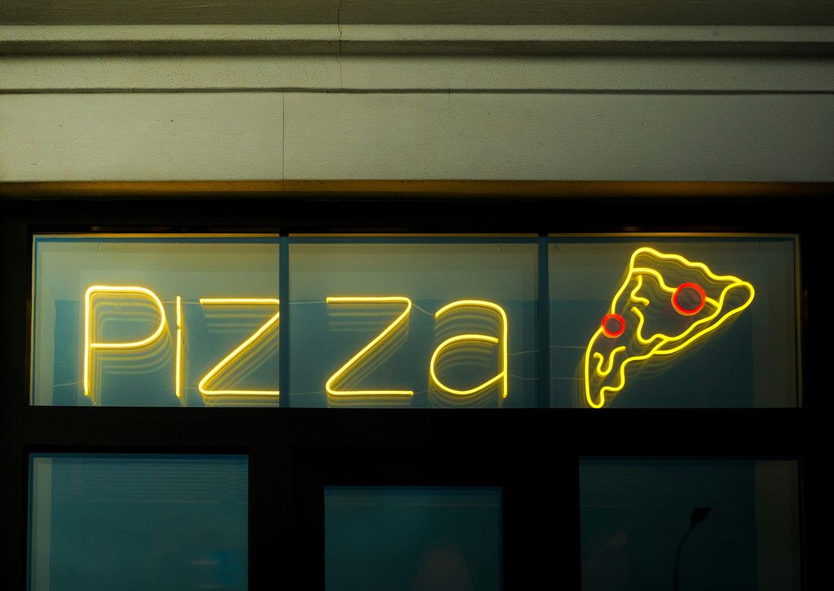 Neon Pizza sign with glowing slice of pepperoni pizza displayed in a storefront window at night.