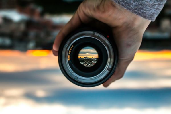 A man holding a camera lens with a city in the background, which you can see through the lens.