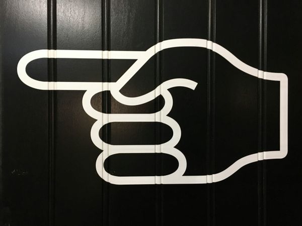 A large white graphic of a hand with the index finger pointing left, displayed on a black paneled wall.