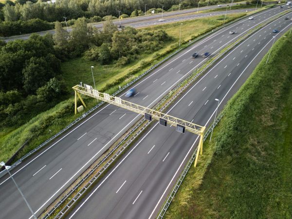 An aerial view of a multi-lane highway with light traffic, bordered by green grass and trees, and a yellow overhead gantry sp