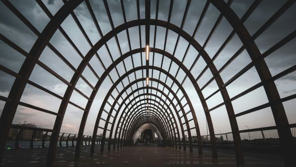 A wide-angle view of a modern arched walkway made of evenly spaced metal beams forming a tunnel-like structure, with a cloudy
