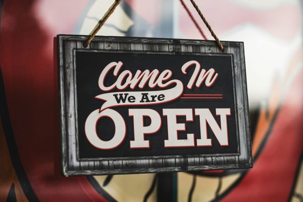 Hanging wooden sign with rope that reads “Come In We Are Open” in bold white and red lettering, set against a blurred storefront background.