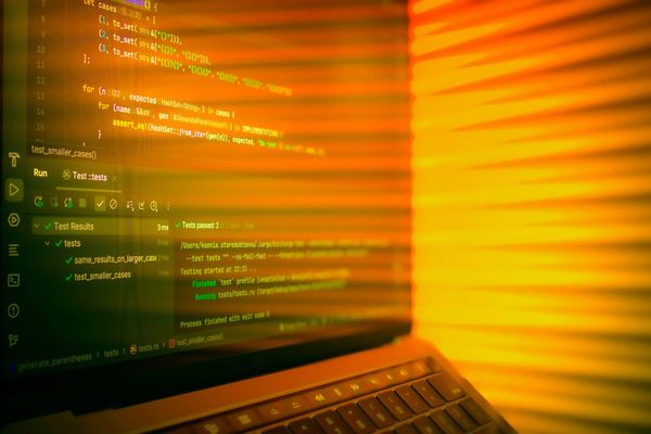 Close-up of a laptop screen displaying code and test results, lit by warm orange sunlight streaming through window blinds.