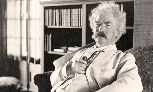 Mark Twain holding a pipe wearing a white suit in a library around 1900.