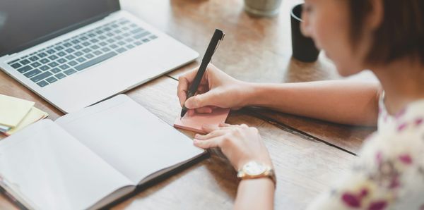 Person writing notes beside an open notebook and laptop.
