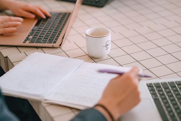Two people working at a table with two laptops, a coffee cup and a notebook in view.