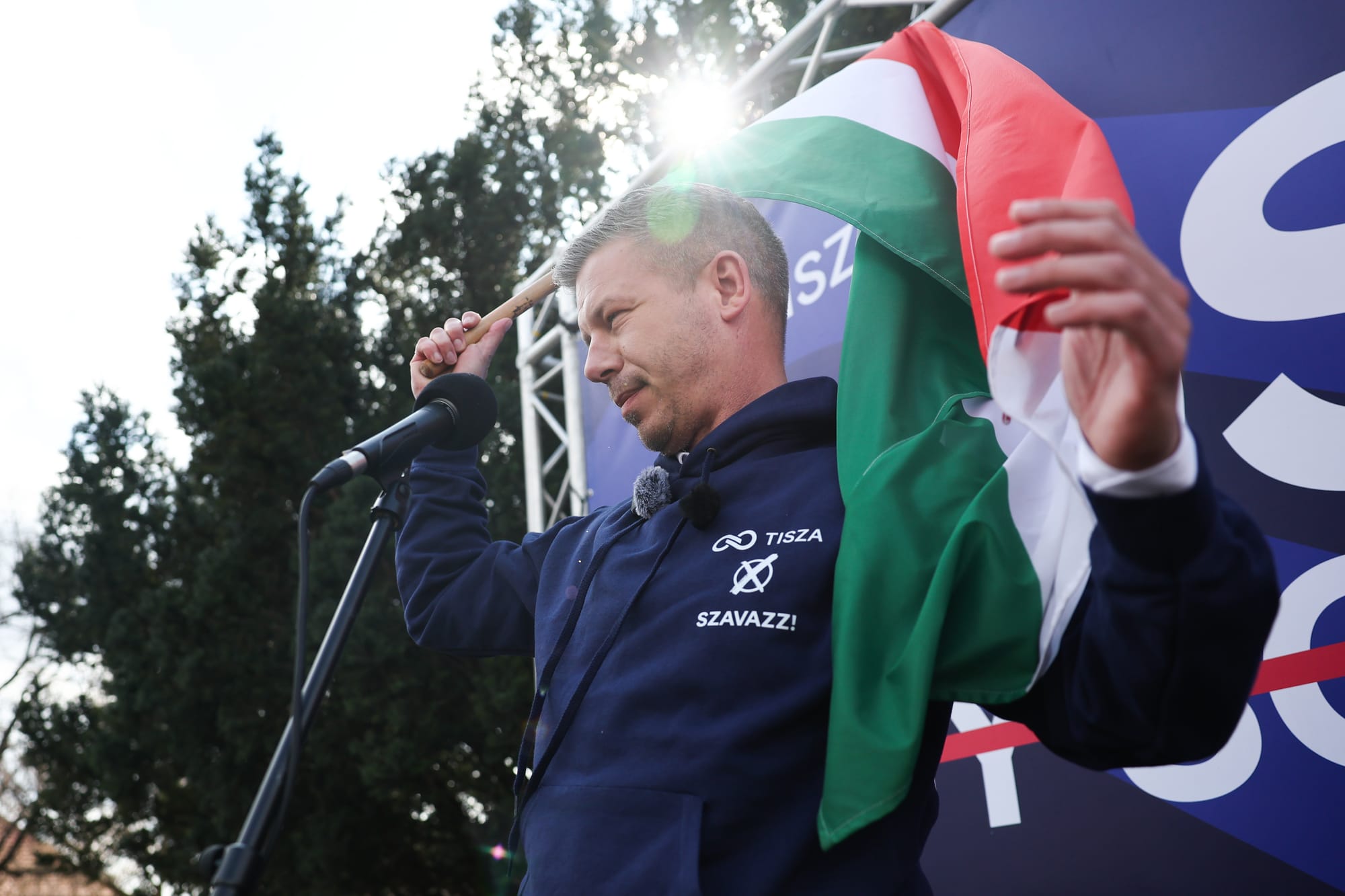 Hungarian politician Peter Magyar stands in front of a mic with a flag in his right hand. he wears a blue sweatshirt with his party's name on left breast