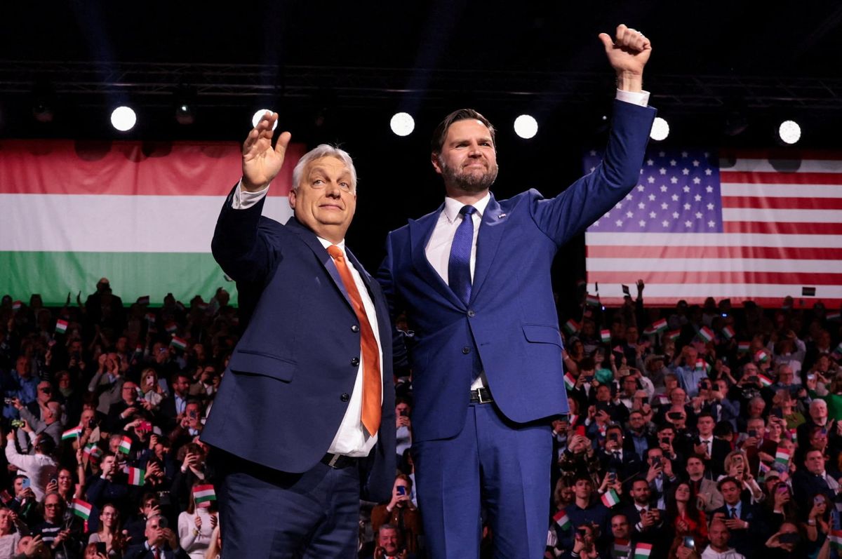 Viktor Orban pictured arm in arm with US vice president JD Vance. Both have a hand raised; Vance gives a thumbs up in a full auditorium with US and Hungarian flags in background.