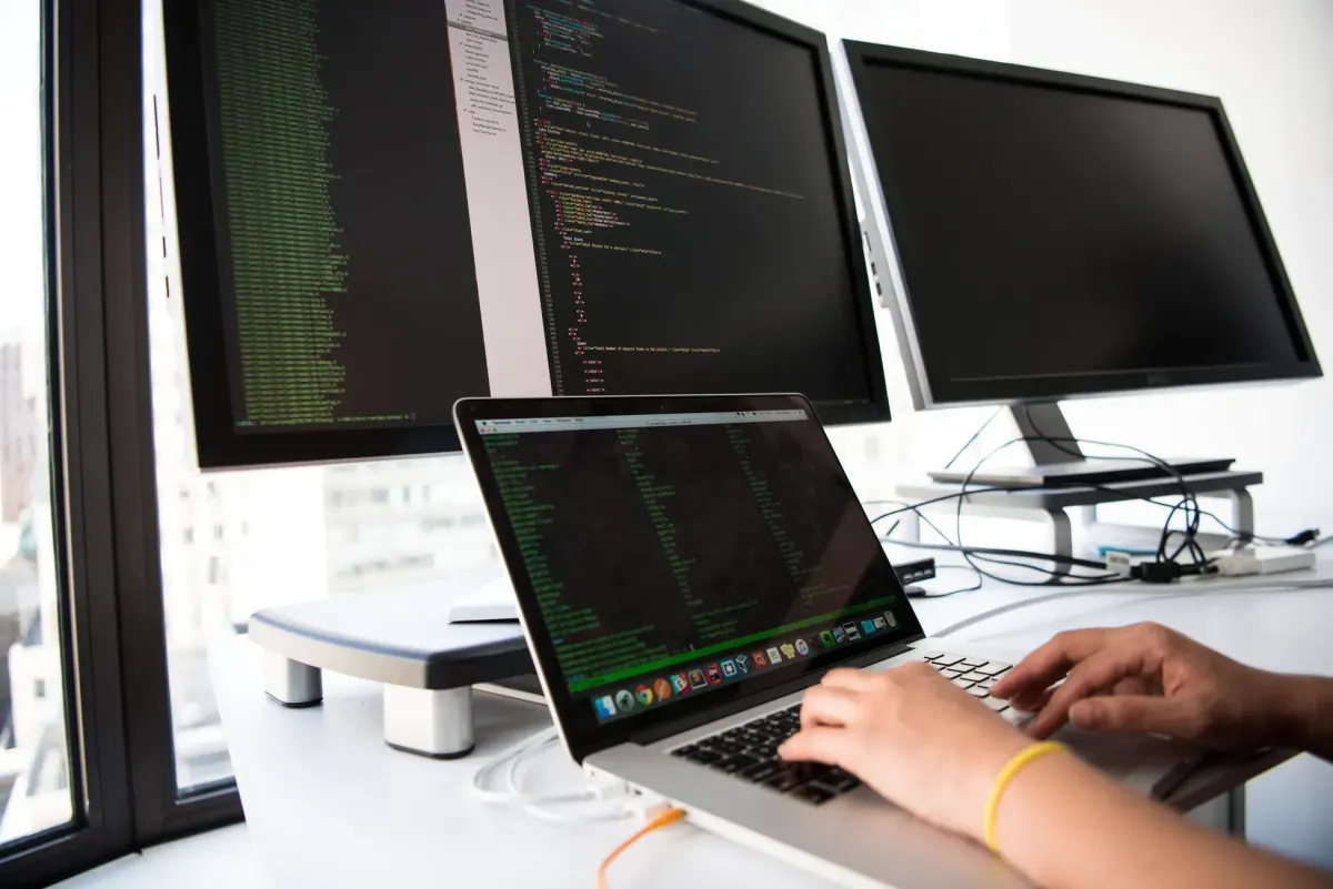 Developer typing on a laptop flanked by two external monitors displaying source code