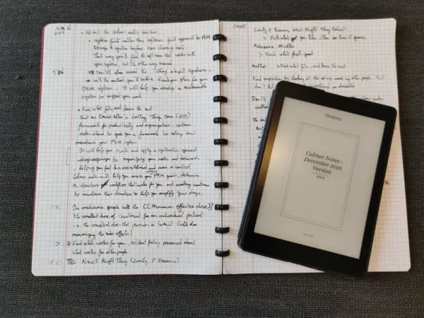 Handwritten notes on graph paper next to a Kindle showing a book cover, reading and taking notes