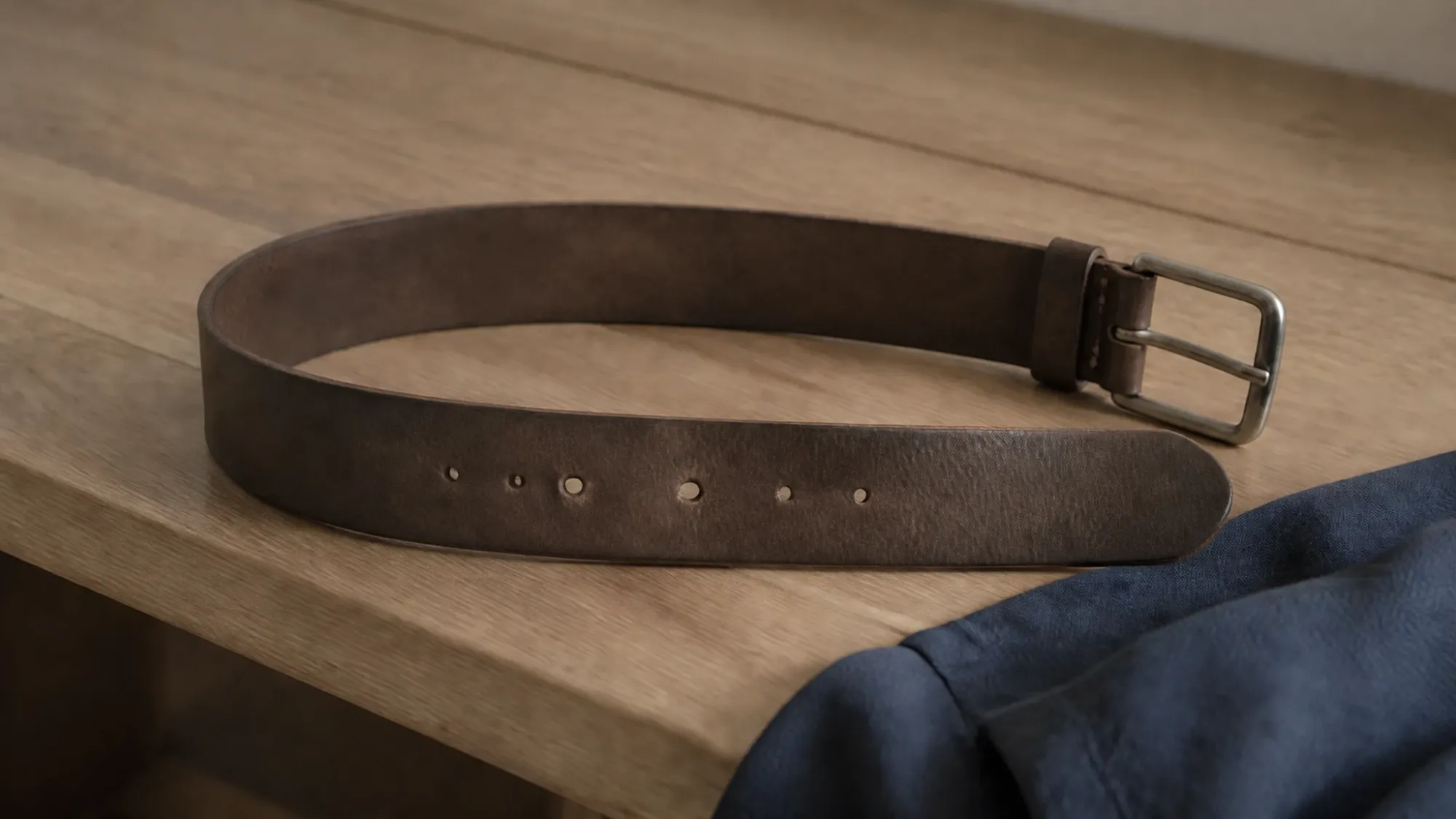 A worn leather belt lying on a pale wooden table, several of its holes visibly stretched from use.