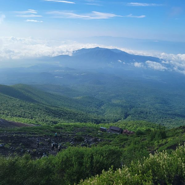 View from the South side of Mt. Fuji located in Japan. A large forest stretches between a valley cutting between the mountain range. Clouds drift near the top of the mountain closest to view.