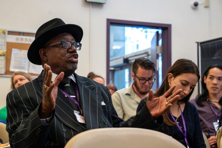 A man in a pinstriped suit sits amid other attendees, explaining to News Relay Network staff.