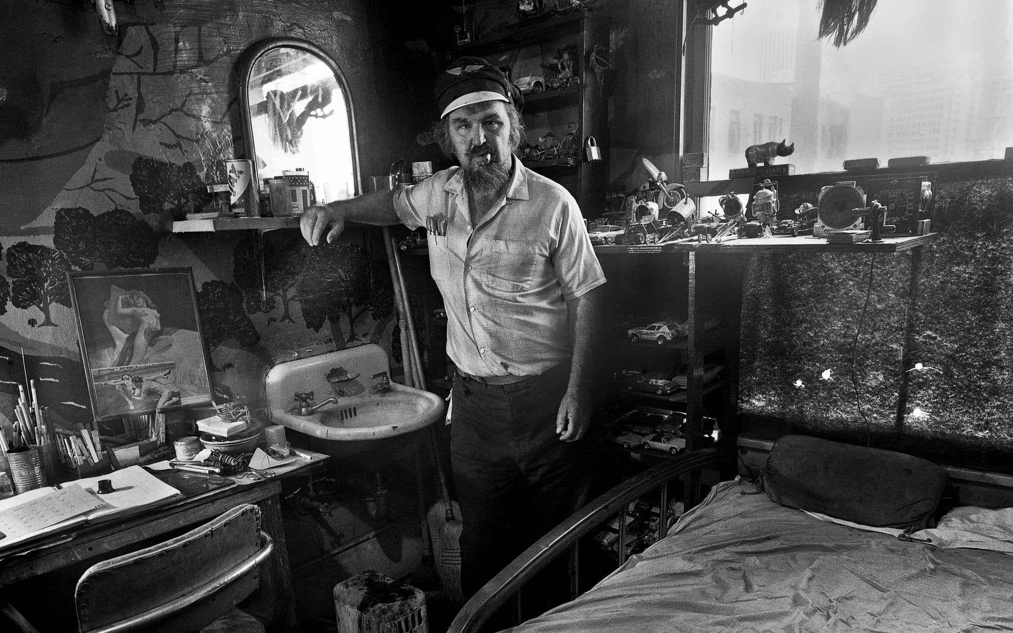 An historic photo of a Skid Row resident in his one-room hotel room with a bed, sink and other possessions, in 1974.