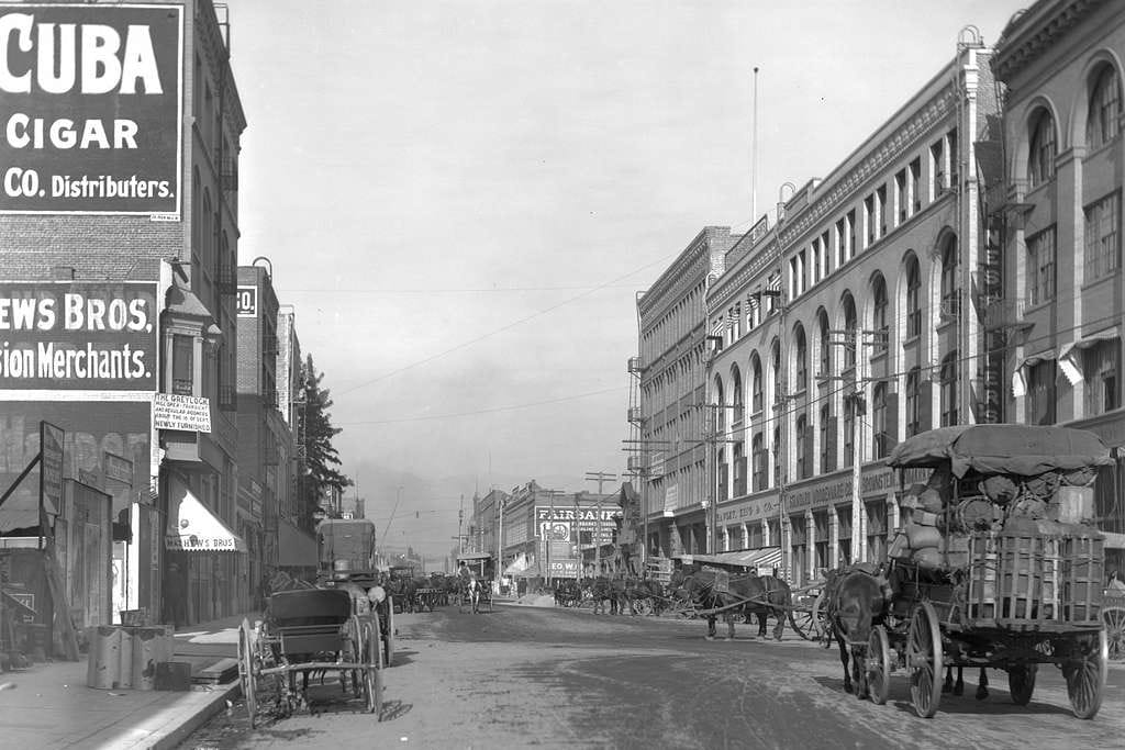 An historic photo of a building-lined street of Skid Row, Los Angeles with horse-drawn carts in 1910.