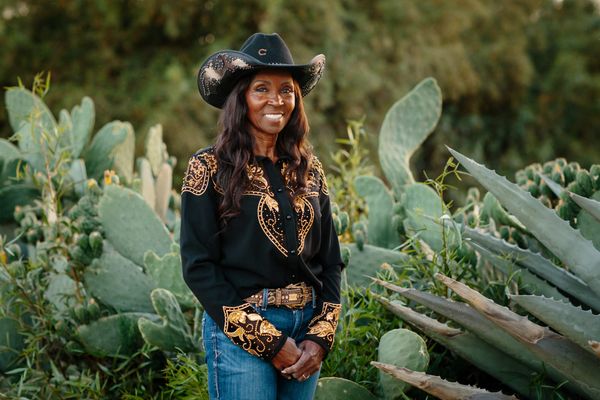 Rodeo pioneer DeBoraha Townson wears a black cowboy hat, shirt and jeans while standing in front of cactuses in her backyard in Agua Dulce, California.