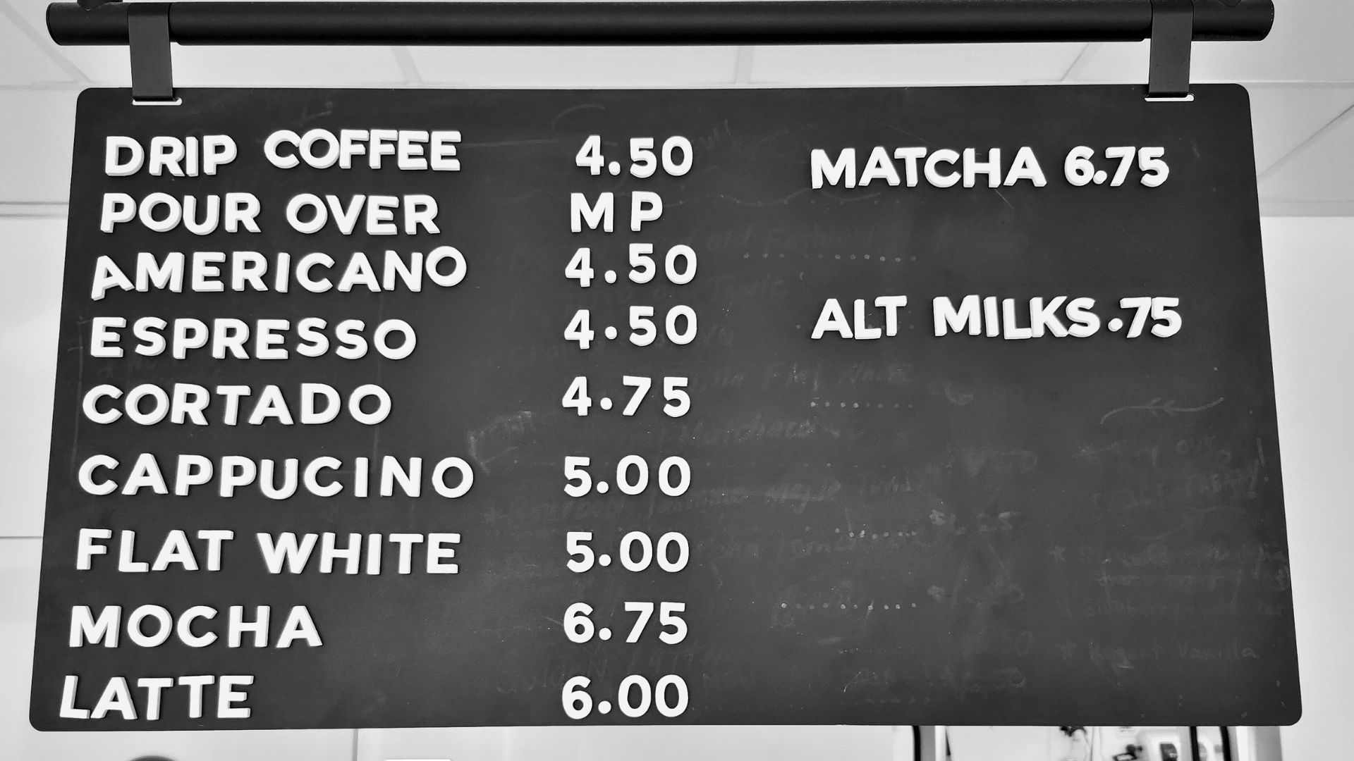 A black and white photo of a hanging menu board at a coffee shop with white block letters on a black eraser board.
