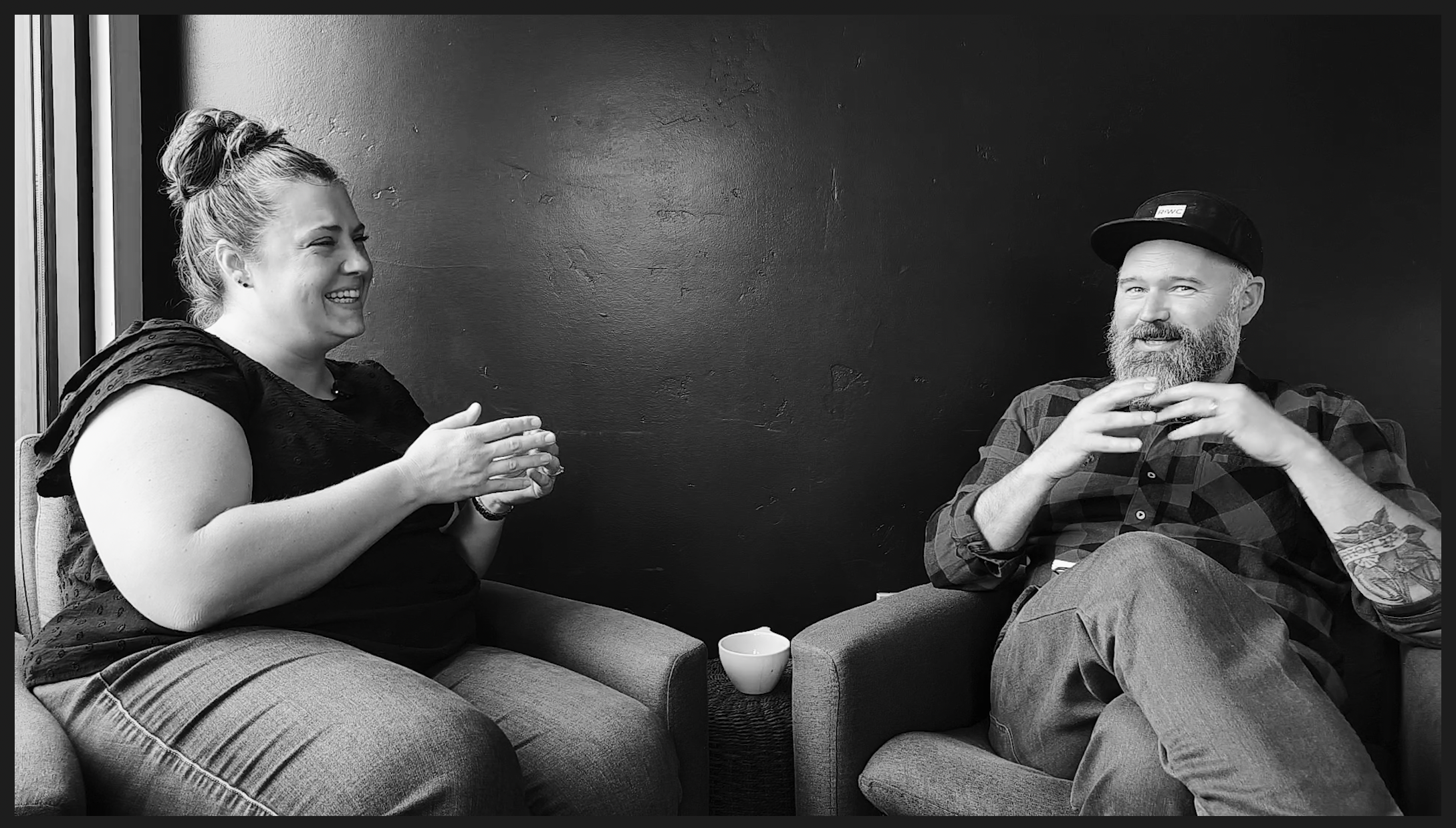A black and white photograph of Jillian Quint (left) and Ryan Woldt (right) sitting in comfortable coffee shop lounge chairs in front of black wall laughing at something that was said during a recording of the Coffee People podcast. A white coffee mug sits on a partially hidden side table between them.