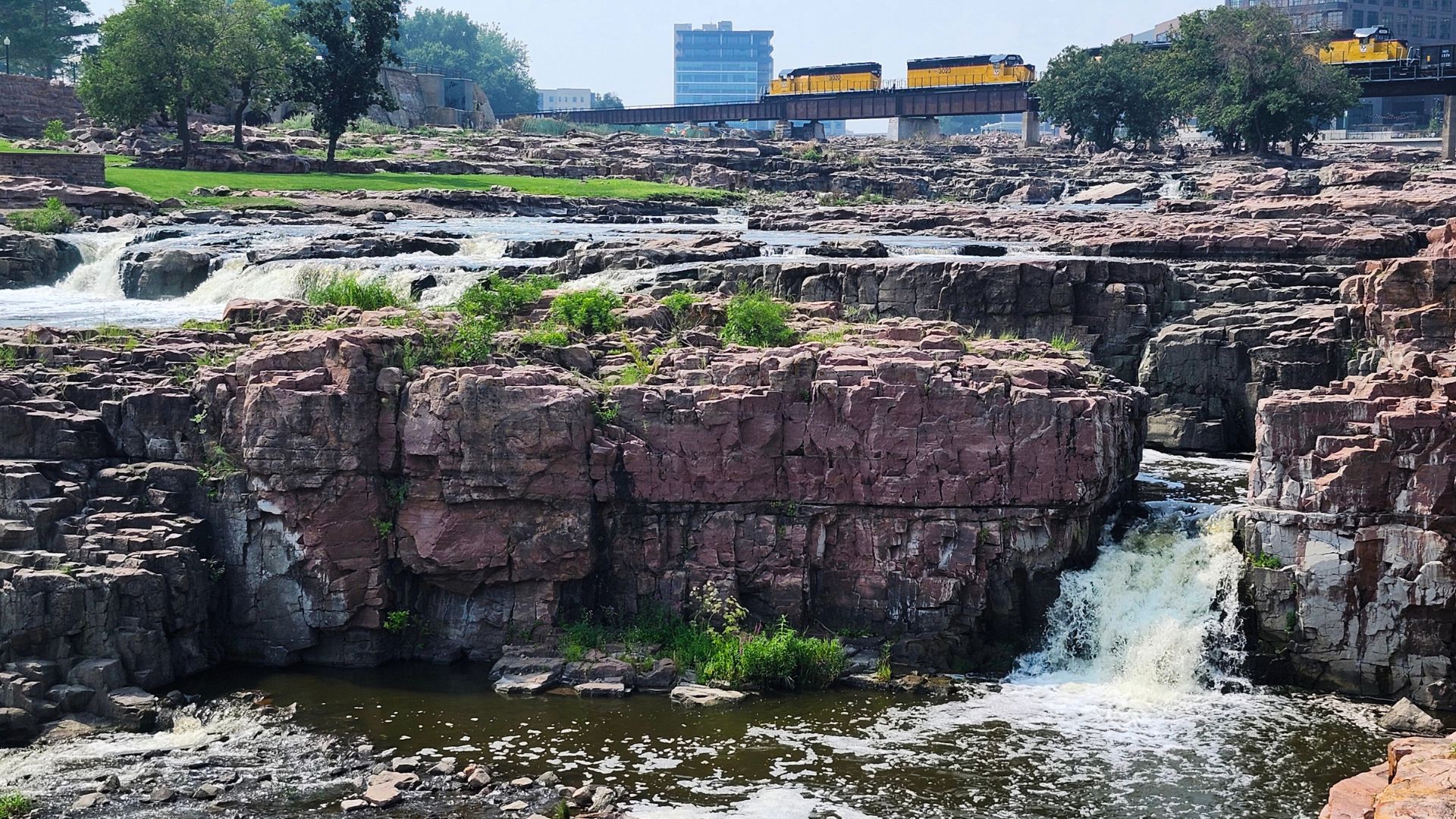 Waterfalls cascade over reddish quartz rock and shrubbery. A yellow and black train crosses over a train bridge in the background in front of Sioux Falls, Downtown skyline.