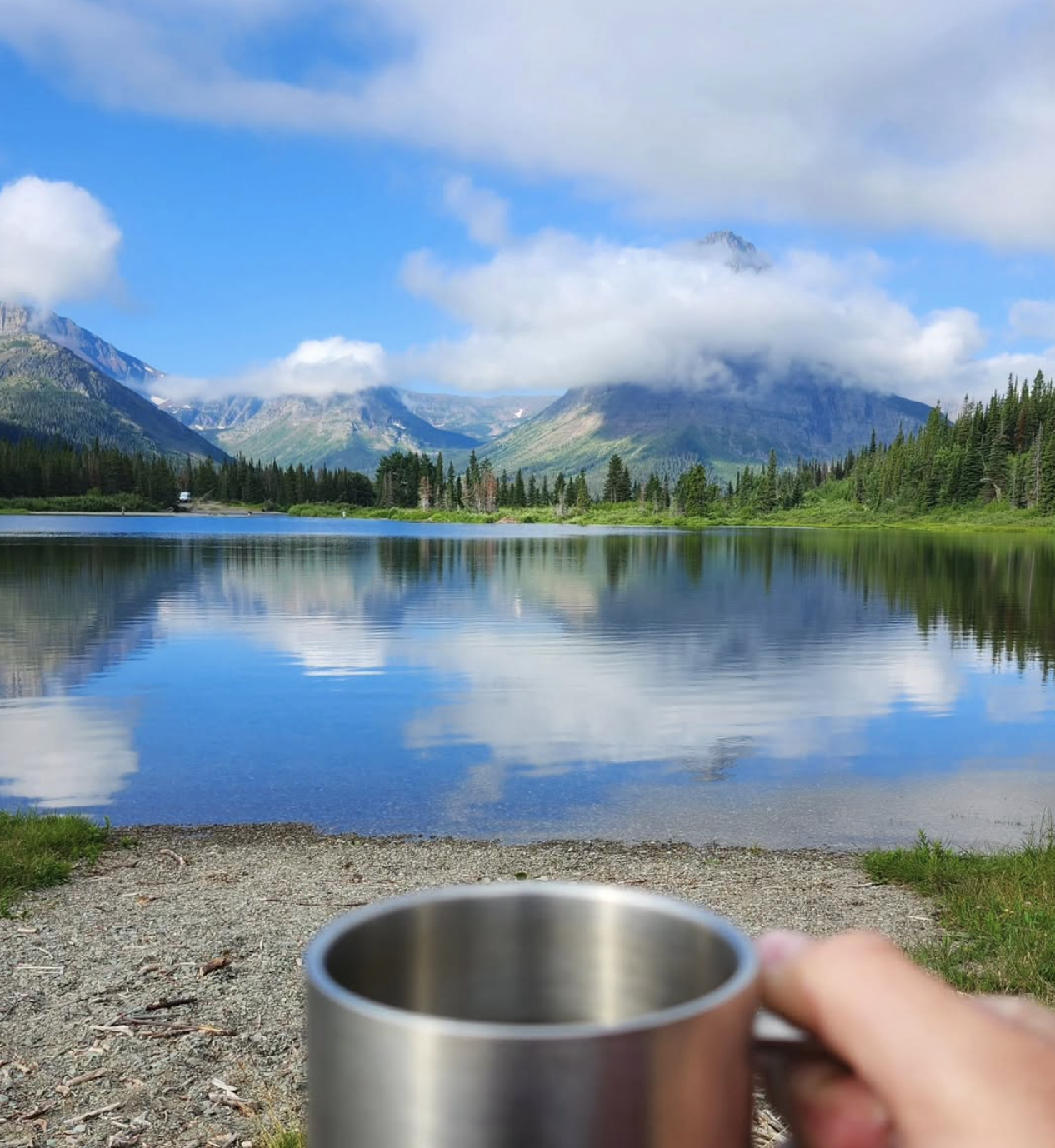 A cup of coffee held up in the forefront of the frame. A mountain partially obscured by a wispy white cloud on a blue sky towers in the background and is reflected on a glassy lake below.