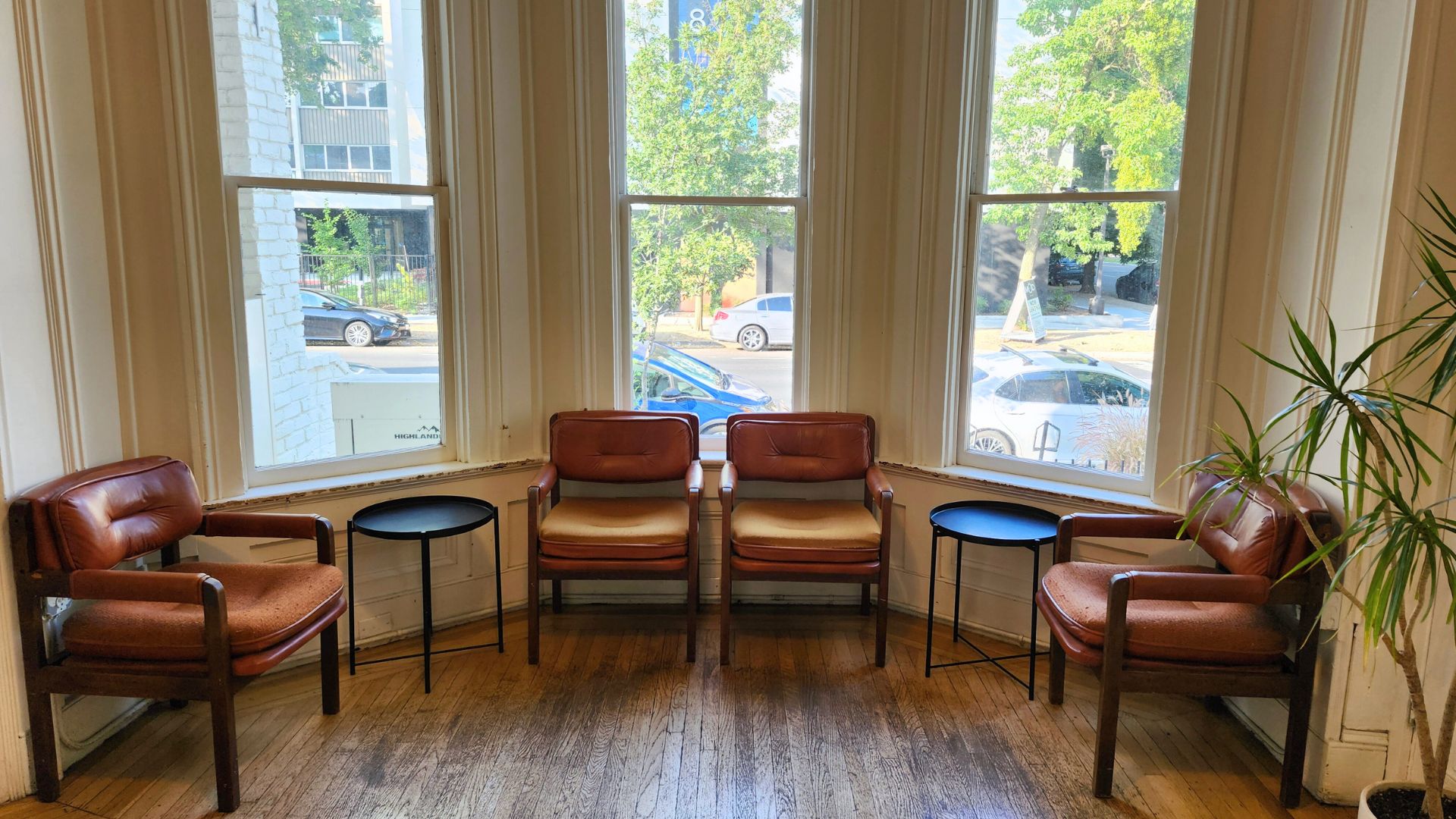 Leather lobby chairs are pushed up against white painted walls between three large vertical windows in a cafe conversation room. The room is a half-round with well-worn wooden floors with narrow boards.