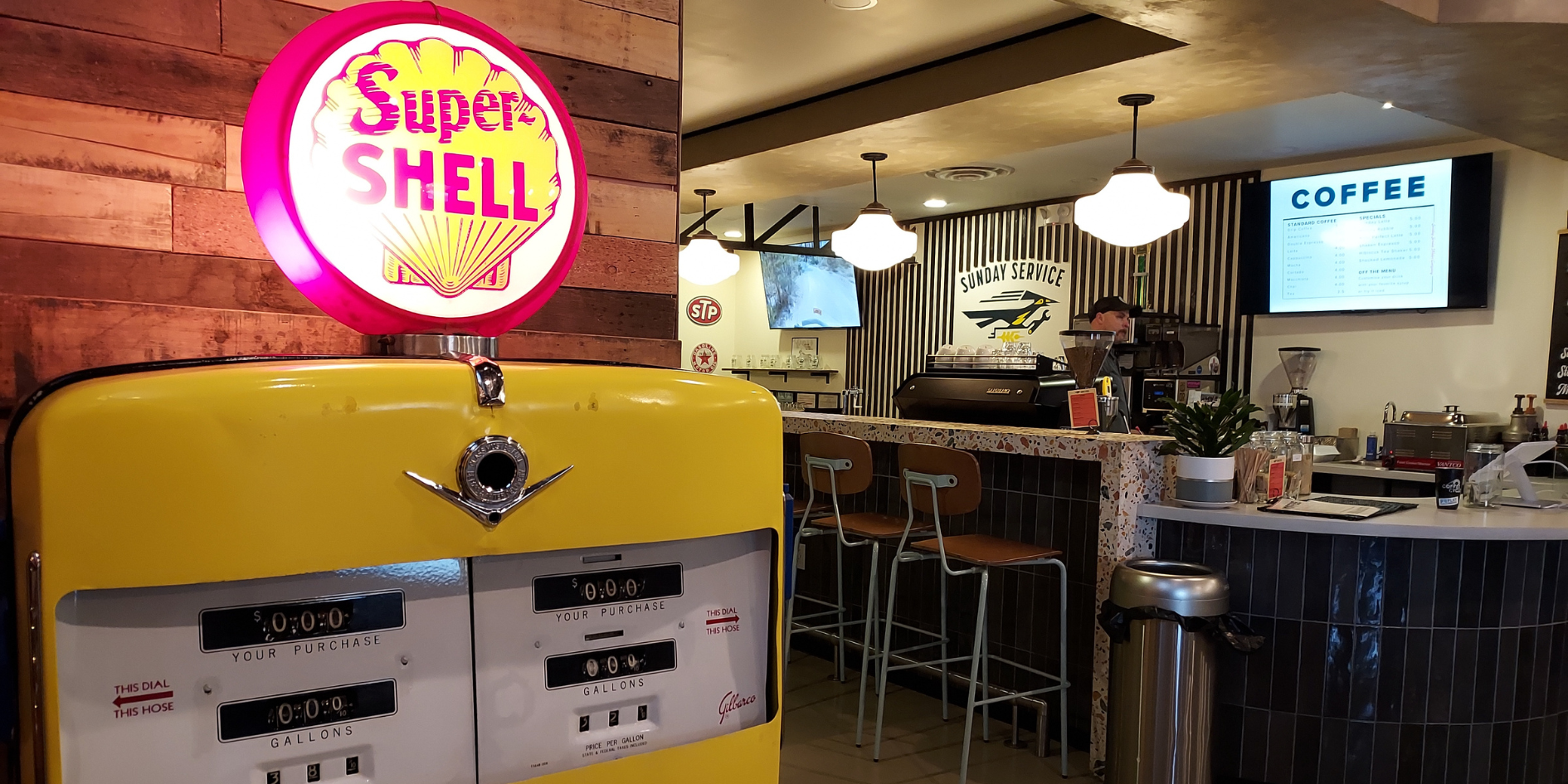 A close-up of a vintage Super Shell gasoline pump is lit up in warm light. To the right, in the background is a barista pulling a shot at an espresso machine behind a tile-faced counter and bar.