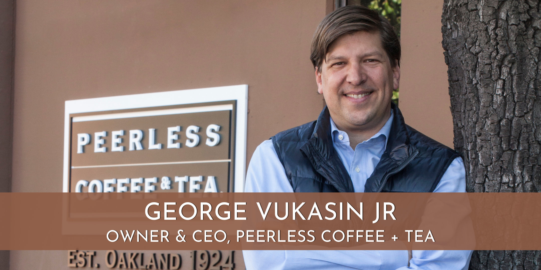A white man smiles at the camera, arms crossed, dark hair brushed to the side. He's wearing a blue collared shirt under a fleece vest, and stands in front of a dark tan wall next to a tree trunk. A sign on the wall says, "Peerless Coffee & Tea. Est. Oakland 1924."