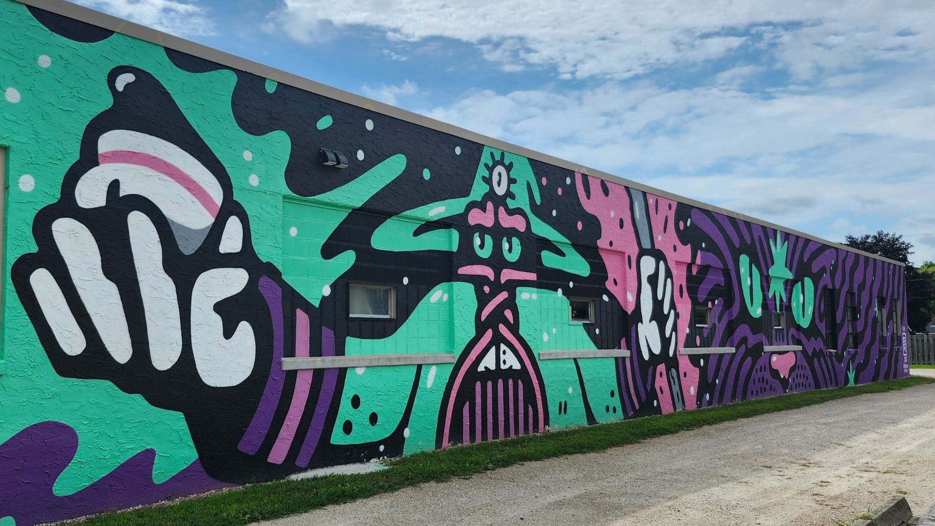 A mural of a wizard holding a coffee cup next to a cartoon cat painted on the side of a long, short brick building. The colors are vibrant seafoam green, pink, white, and purple against a black background.