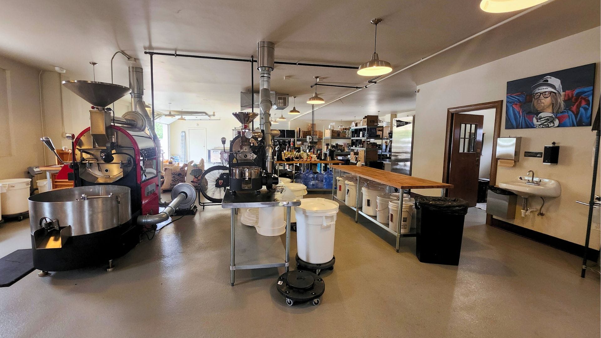 A wide shot of a coffee roasting space. From left there are two mid-sized drum coffee roasting machines lined up. Buckets of green coffee are stored under long tables, and on the right wall a painting of Dana Carvy as Garth from Wayne's World is mounted above a handwashing sink.