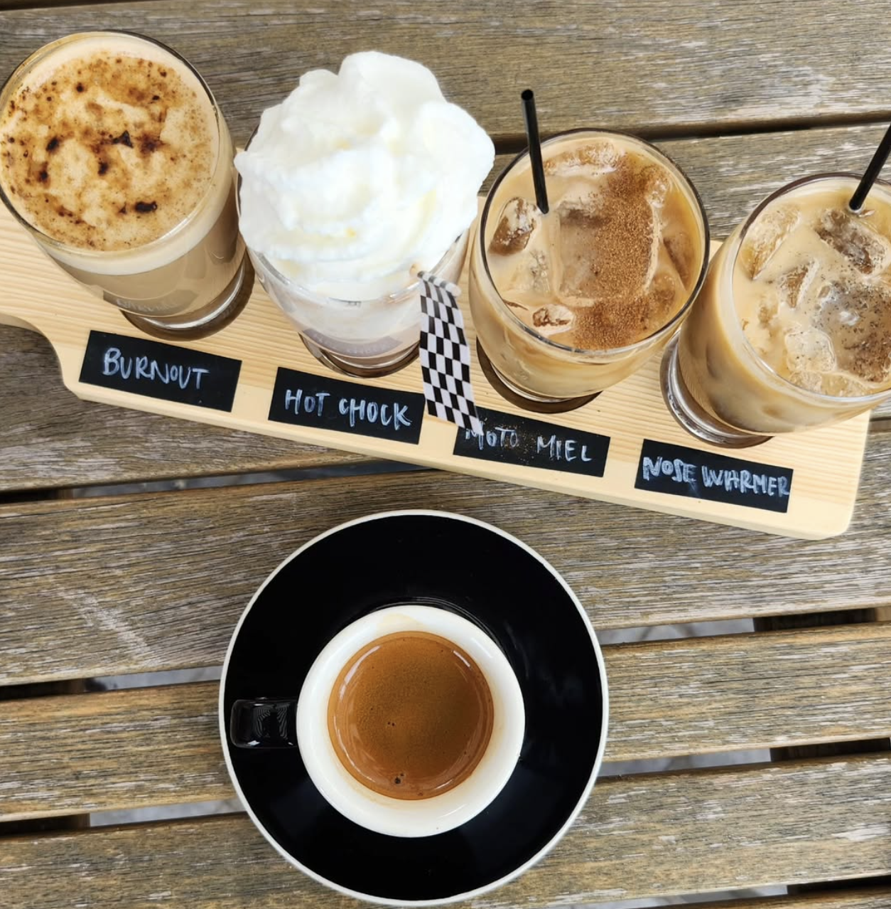 A top view of a flight of four coffee drinks on a wooden paddle at the top of the image and an espresso in a black cup and saucer with white trim. The glassware sits on a weather worn patio table.