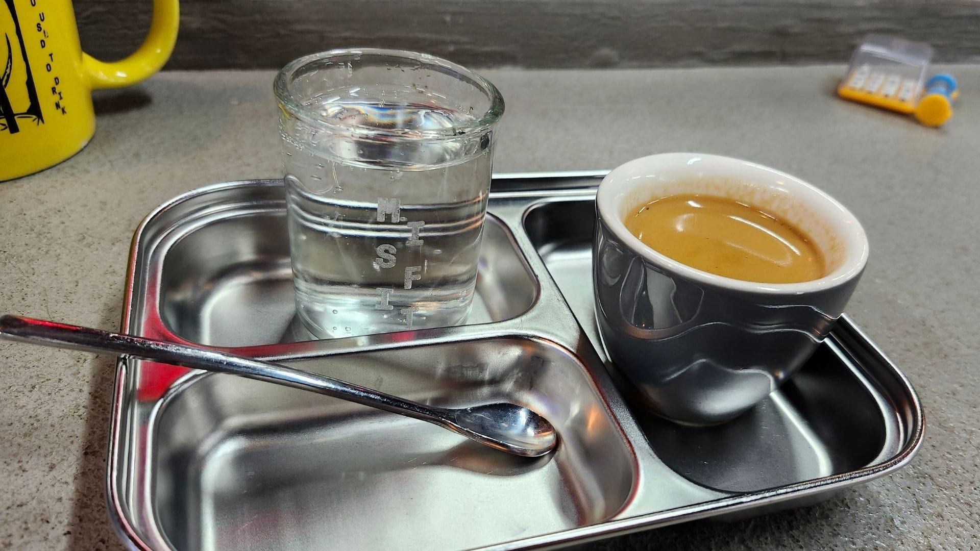 An espresso on a tin with a small spoon, and a short glass filled with seltzer.