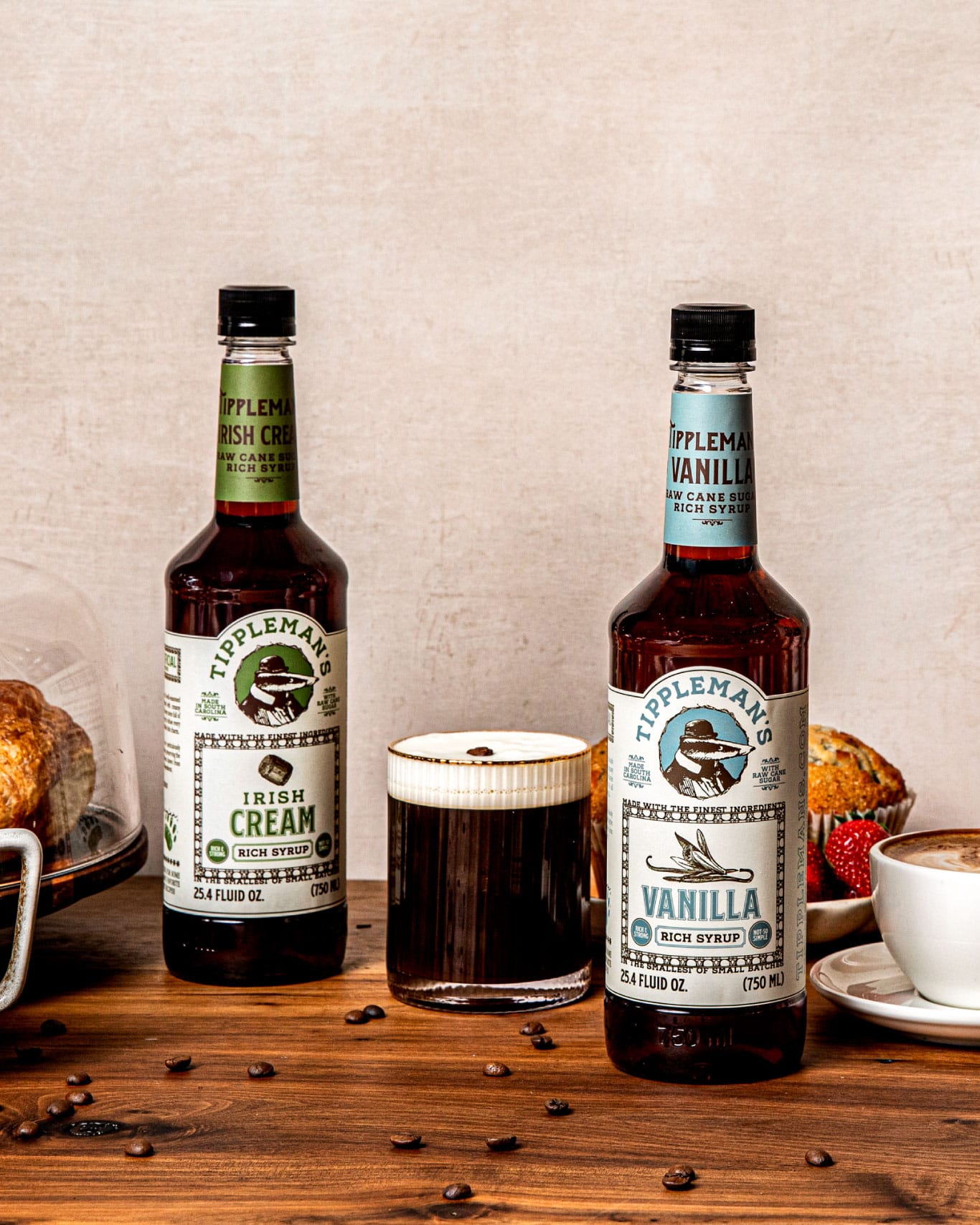 Tall bottles of Tippleman's coffee syrups frame a coffee in crystal glass. The coffee is dark colored with thick, creamy head, and a scattering of coffee beans on the wood counter around it.
