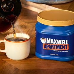 Coffee is poured into a white coffee mug on a wood table next to a blue plastic bin of coffee with a light brown lid. A branding label says "Maxwell Apartment" with a photo of the same mug.