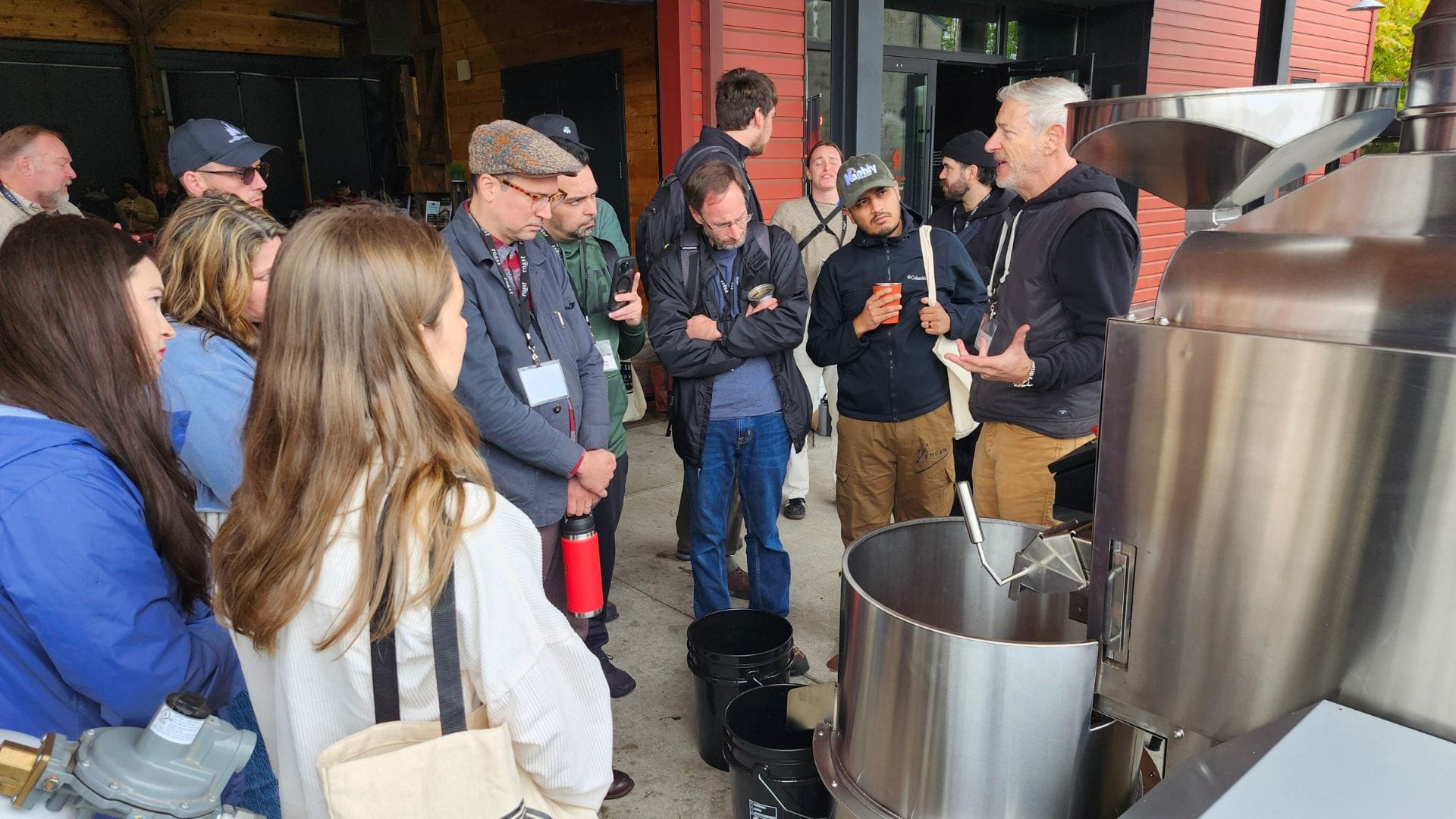 A group gathers around a stainless steel roaster to watch a coffee roasting demonstration outside of a brick red building.