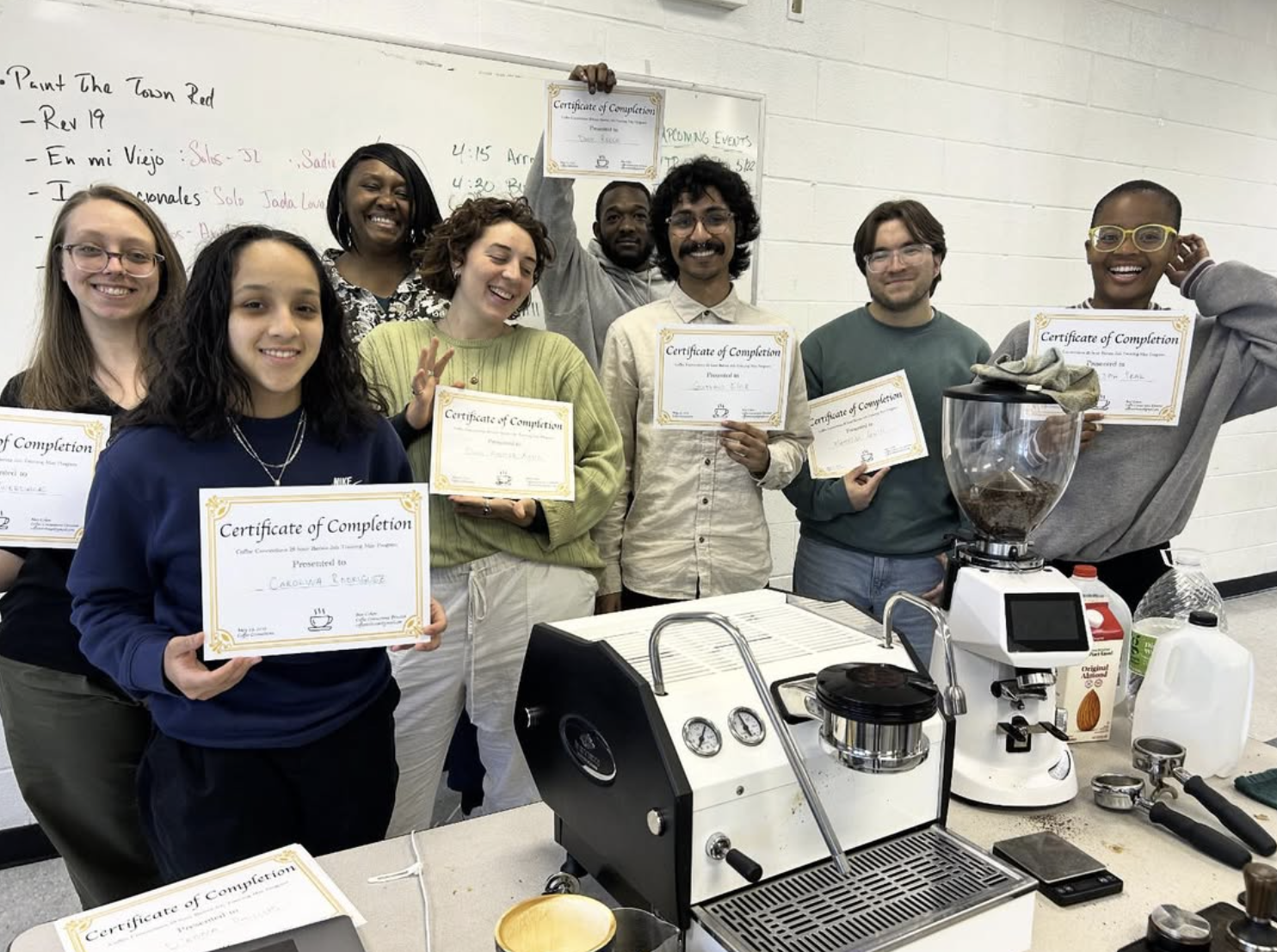 A group of recent barista skills training graduates hold certificates up to the camera while in front of a white board in a classroom and behind a table filled with coffee equipment including a chrome and black espresso machine.