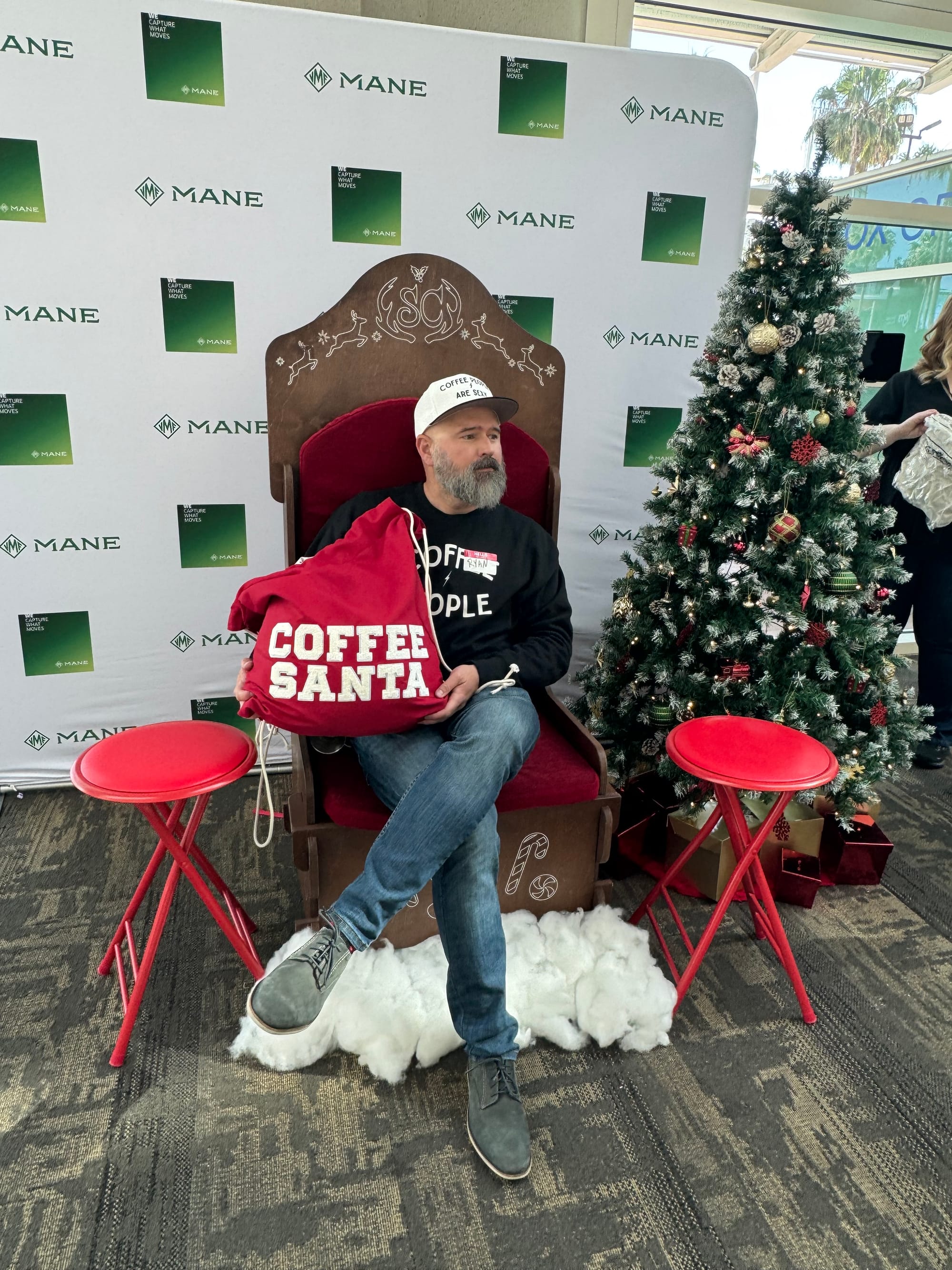 A salt-and-pepper bearded man holding a red Santa sack that says "Coffee Santa" sits on a holiday themed chair next to a Christmas Tree.