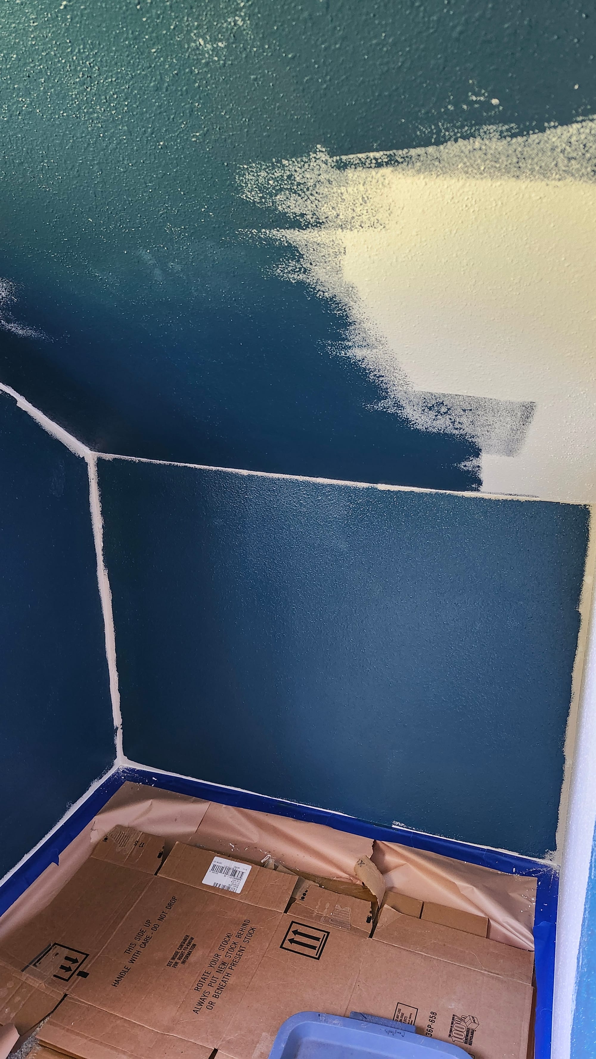 A closet is being painted. Dark blue paint covers most of the beige. The trim is covered with blue painter's tape and the floor covered by cardboard and paper to prevent spills.