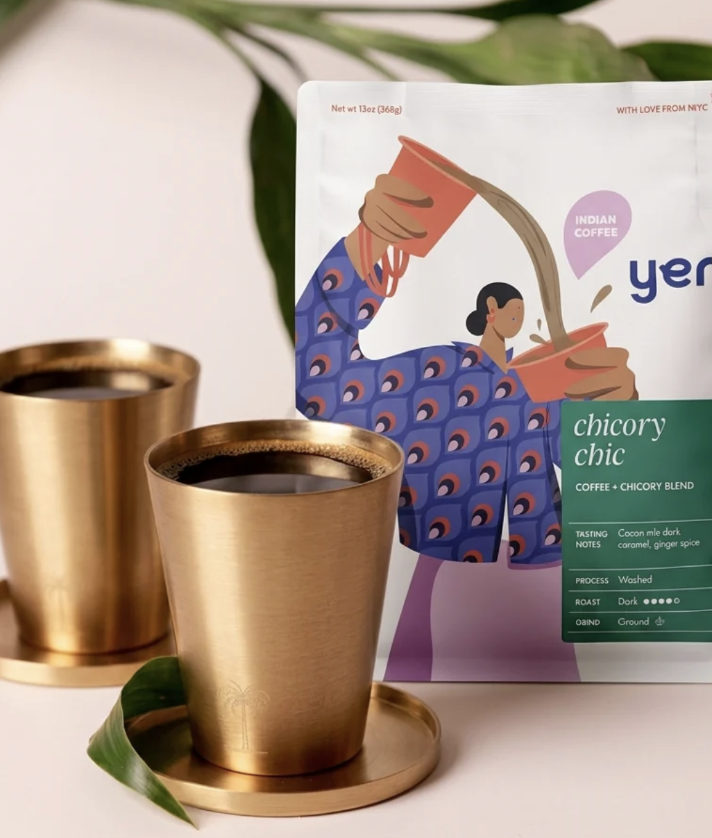 Close up of coffee in two brass coffee cups in front of the package which features an artistic, illustrated woman pouring coffee into a cup by tipping the pot from high over head.
