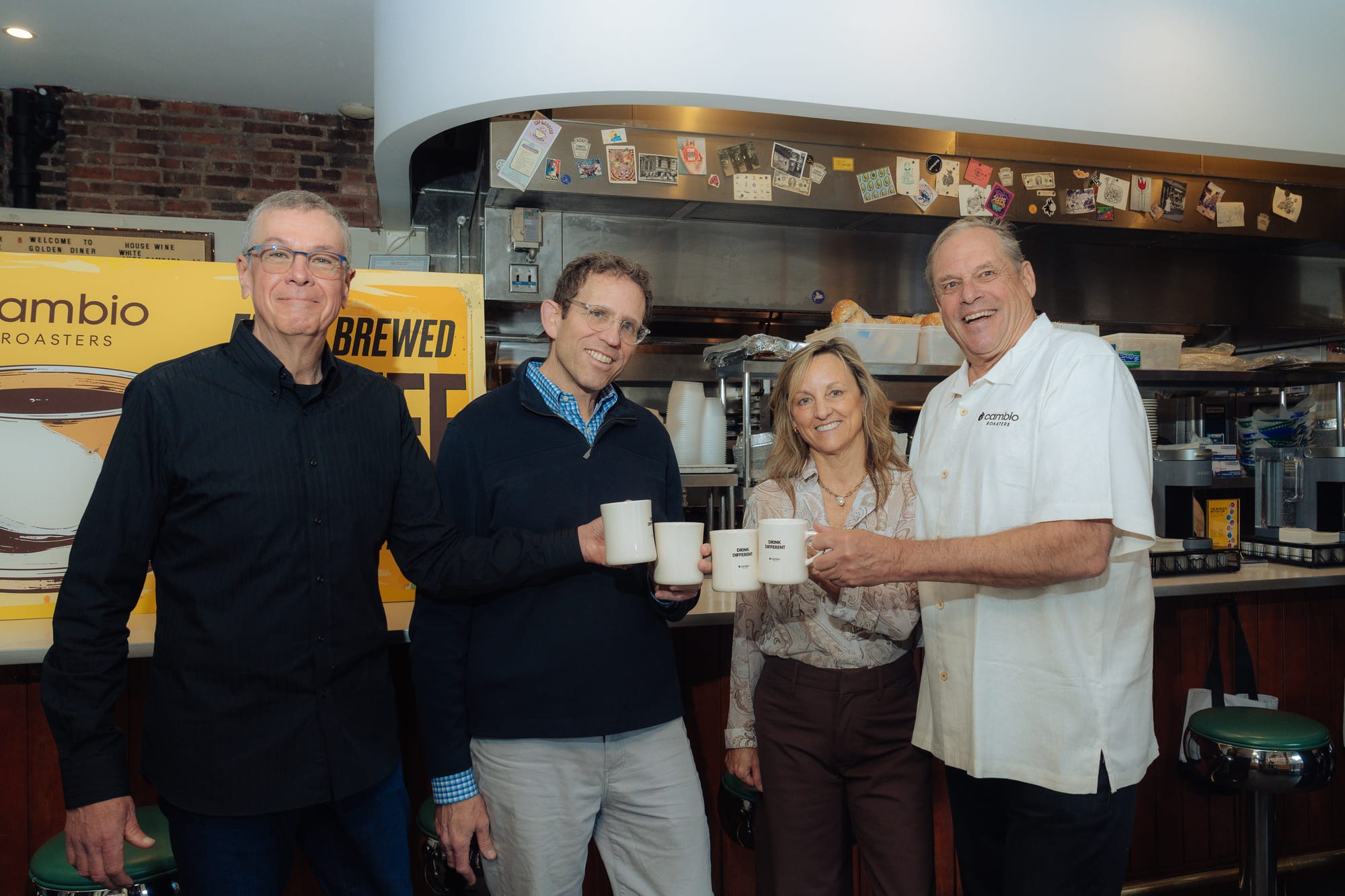Four people "cheers" their coffee mugs while smiling for the camera. From left: A man in a black button down with short cropped salt-and-pepper hair wearing wire-rim glasses. A bookish looking younger man in a blue sweater over a checked button down. A woman in brown slacks and a light beige buttoned blouse with dusty blonde hair, and a taller man with a big grin, white hair combed over and a white bowling team-style button down with the Cambio Roasters logo featured over the heart. They're standing in front of bar and kitchen in a classic coffee shop diner.